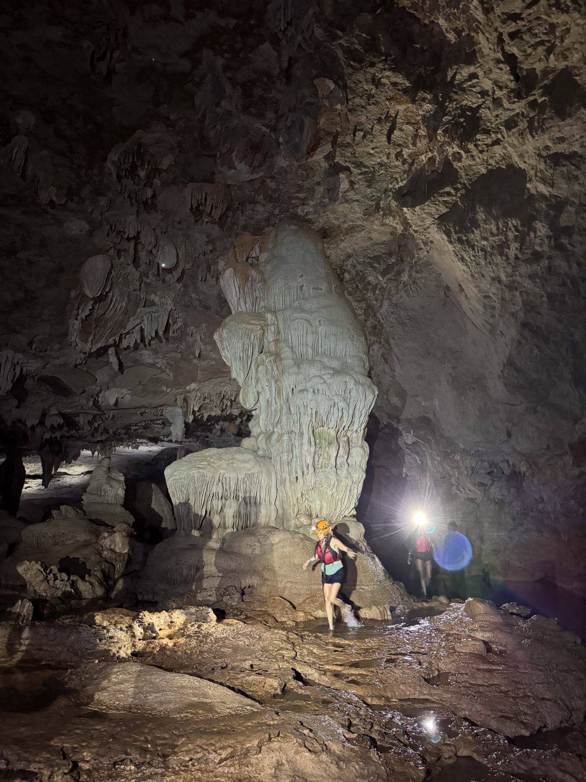 A student in a helmet and headlamp wading past a large limestone formation inside a cave