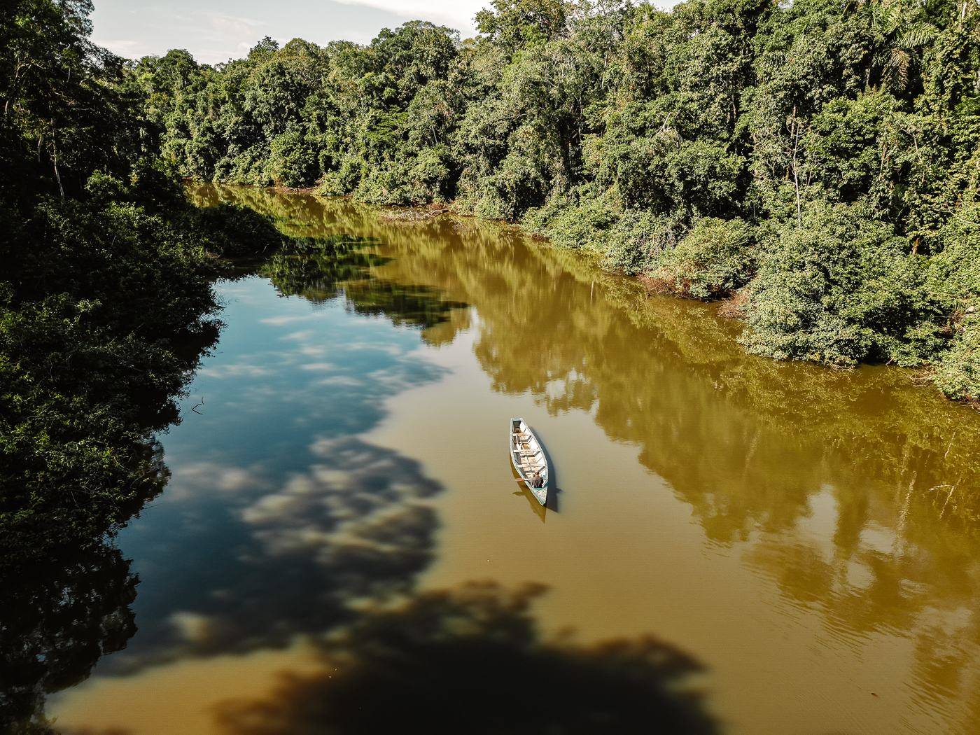 Aerial view of a wooden canoe on a brown oxbow lake, with rainforest reflected in the water
