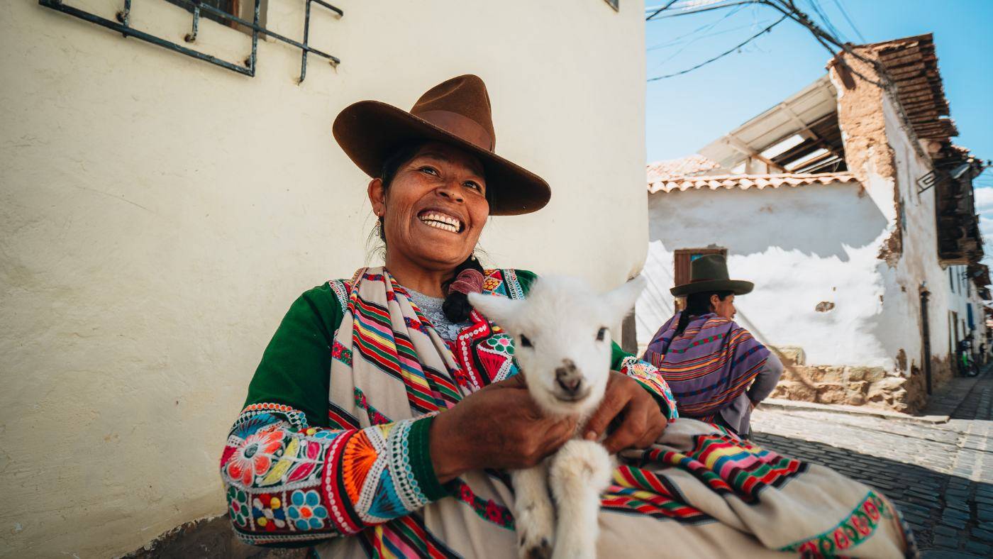 A smiling Quechua woman in an embroidered shawl holding a young lamb on a Cusco street