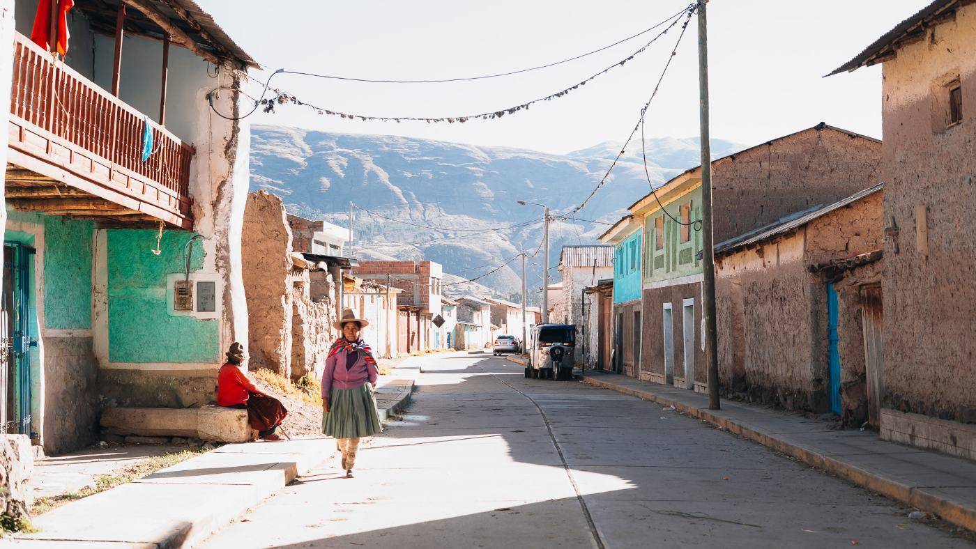 A woman in a pleated skirt walking down a sunlit Andean village street