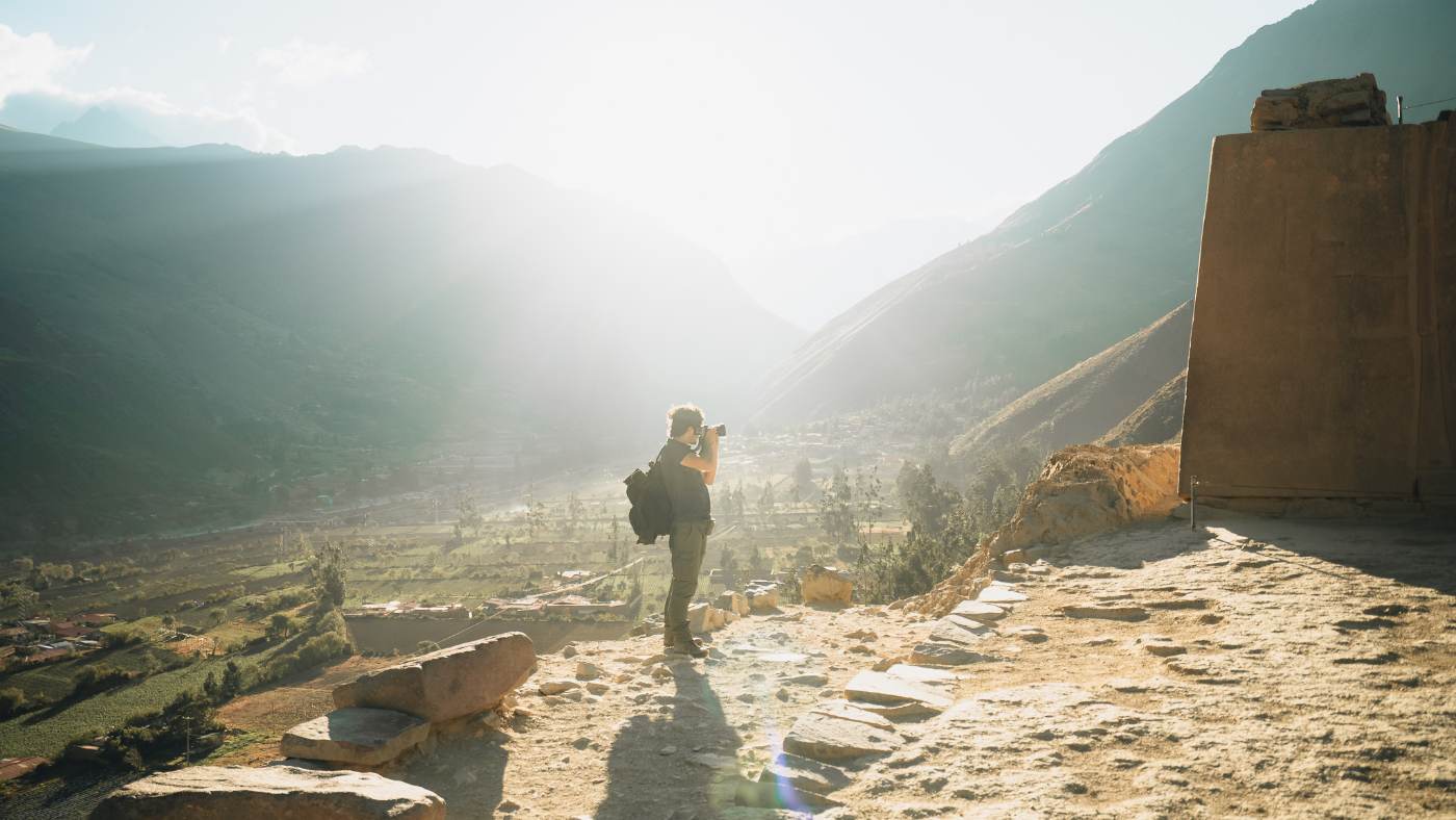 A traveler photographing a sunlit Andean valley from a stone overlook